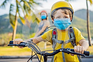 Active school kid boy and his mom in medical mask and safety helmet riding a bike with backpack on sunny day. Happy