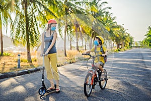 Active school kid boy and his mom in medical mask and safety helmet riding a bike with backpack on sunny day. Happy