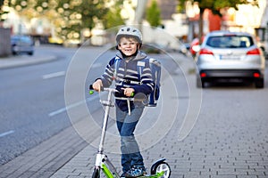 Active school kid boy in safety helmet riding with his scooter in the city with backpack on sunny day. Happy child in