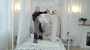 Active happy senior Caucasian grandparents couple jumping, having pillow fight on bed at home