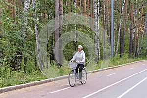 Active senior caucasian woman with gray hair is riding bike on bicycle path in forest park.