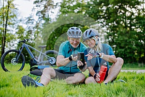 Active senior couple resting after bicycle ride at summer park, sitting on grass and using smartphone.