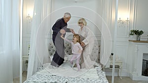 Active senior grandparents couple in pajamas jumping with granddaughter child girl on bed at home