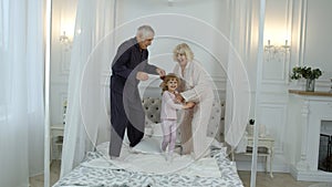 Active senior grandparents couple in pajamas jumping with granddaughter child girl on bed at home