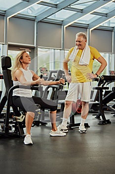 Active senior man, with towel on shoulders, talks with woman doing exercises lifting weights at modern gym.