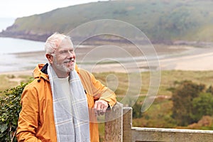 Active Senior Man Walking Along Coastal Path In Fall Or Winter By Gate With Beach And Cliffs Behind