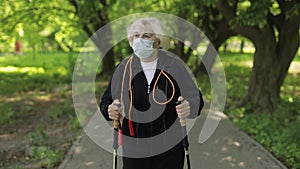 Active senior old woman in mask training Nordic walking in park at quarantine