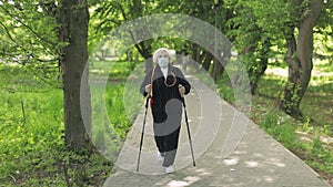 Active senior old woman in mask training Nordic walking in park at quarantine