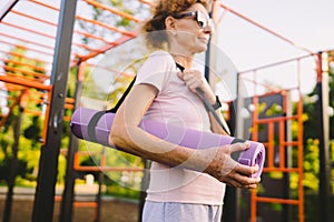 Active senior woman with fitness mat and water bottle in hands walking on workout outdoor gym. Happy mature lady