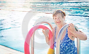 Active senior woman (over the age of 50) in sport goggles, swimsuit, with swim noodles on ladder handrails.