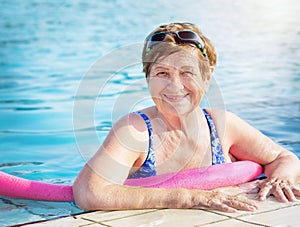 Active senior woman (over age of 50) in sport goggles, swimsuit and with swim noodles in swimming pool.