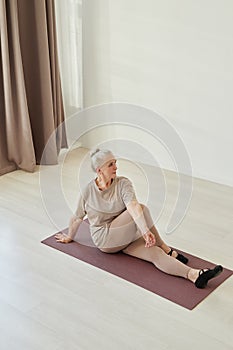 Active senior woman in sports clothes sitting on mat during physical training