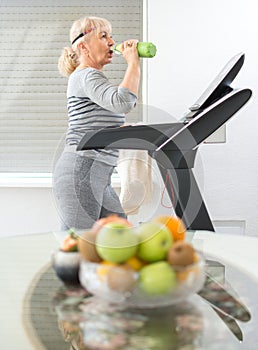 Active senior woman in sportswear drinking water while walking on a treadmill at home