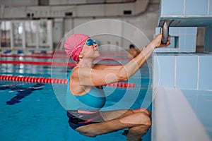Active senior woman swimmer holding onto starting block preparing to swim in indoors swimming pool.