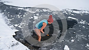 Active senior woman in swimsuit breaking ice with axe outdoors in winter, cold therapy concept.