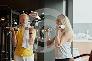 Active seniors, man and woman, doing strength training for biceps with dumbbells with smiles in modern fitness center.