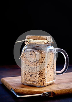 Active sourdough starter in glass jar. Rye leaven for bread on wooden cutting board on black background. Close-up. Dark low key