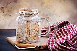 Active sourdough starter in glass jar. Rye leaven for bread on wooden cutting board and kitchen towel on brown background. Close-