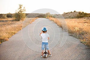 Active sport little child in helmet and denim shorts standing on desert road with scooter on summer warm sunset
