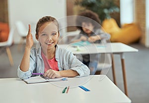 Active student. Cute little school girl smiling at camera raised her hand while sitting at the desk in classroom