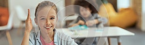 Active student. Cute little school girl smiling at camera raised her hand while sitting at the desk in classroom