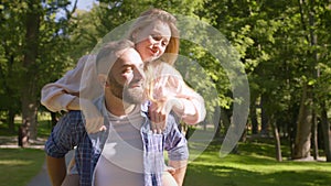 Active woman jumping on loving boyfriend back, happy man riding her in green summer park, slow motion, tracking shot