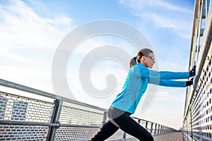 Active woman with sunglasses doing leg stretching on a bridge, hodling onto fence