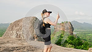 Active woman treks down mountain trail, Sigiriya Rock in view. Hiker with backpack captures lush scenery on smartphone