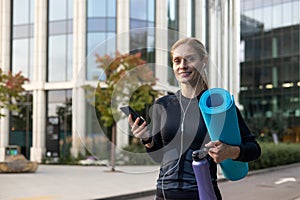Active woman with yoga mat and water bottle using smartphone in city