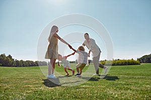 Active young parents spending time together, holding hands in circle with their two little kids, boy and girl in green