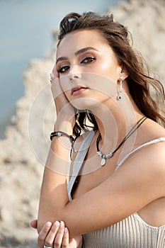 Active young woman sits on beach in relaxed pose and adjusts her hair with hand, blurred background