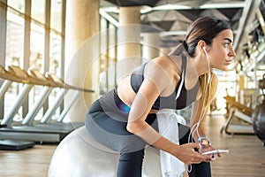 Active young woman sitting on fitness ball and listening music with smart phone in fitness training gym.