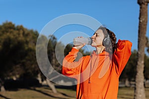 Active young woman in wireless headphones drinks water from bottle after exercising outdoors at park. Female athlete in