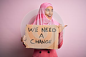 Activist woman wearing pink muslim hijab holding banner with we need a change message pointing and showing with thumb up to the