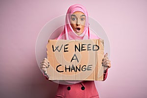 Activist woman wearing pink muslim hijab holding banner with we need a change message scared in shock with a surprise face, afraid