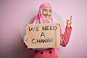 Activist woman wearing pink muslim hijab holding banner with we need a change message surprised with an idea or question pointing