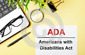 ADA Americans with Disabilities Act is written in red on a white piece of paper on a light yellow background next to a laptop, pen