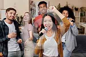 Add music to your party. Young pretty asian woman holding microphone and singing while having fun with friends at home