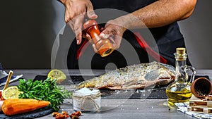 Add pepper to river carp with spices in chef hands, dark background. Food recipe photo, copy text