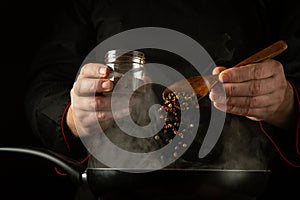 Adding pepper to a hot frying pan with a spoon in the cook hand. Concept on a dark background of preparing a delicious lunch in