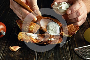 Adding sour cream with a spoon to potato pancakes on the kitchen table. Low key concept of preparing a delicious dish for lunch in