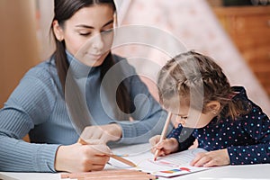Adirable little girl with her beautiful young mom draws in the album using color pensins. Cute girl paints coloring page