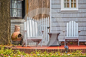 Adirondack chairs and chiminea and wicker table on patio under stree in front of house with grey wooden siding and picket fence