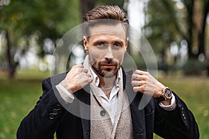 Adjusting the collar of a dark trench coat handsome man standing outdoors in an autumn park smiling looking at the