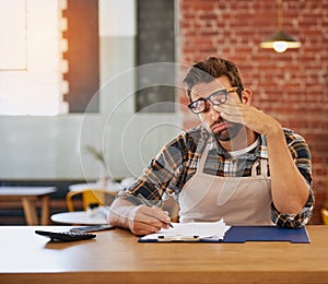 This admin will be the end of me. Shot of an exhausted young business owner doing admin at a table in his coffee shop.