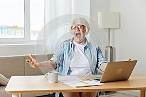 an admiring, happy elderly woman is sitting at her desk working on a laptop and spreading her hand to the side shouts