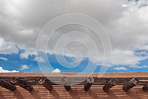 Adobe roofline with exposed vigas and open latillas, blue sky with clouds