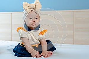 Adolable Asian newborn baby girl, big eyes, puffy cheeks, cutely dressed in yellow and blue, sitting on a white bed looking at
