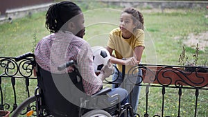 Adolescent African American girl talking in slow motion with disabled man in wheelchair playing soccer ball. Portrait of