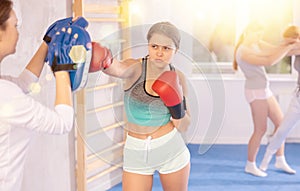 Adolescent girl in boxing gloves punching pads in hands of coach in gym